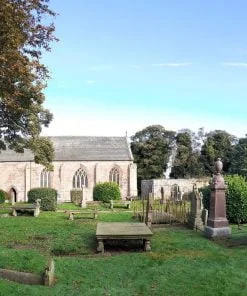 A graveyard with many monuments and gravestones occupies the foreground. Two large deciduous trees are to the left. Centre background is the sidewall of a smallish church building dating from the 14th century. The door is to the left hand end, and three gothic arched windows occupy the rest of the wall. There are bushes and trees behind it, and a blue sky above.