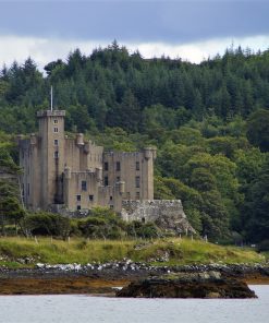 The grey harling towers of Dunvegan Castle are central in this picture. The picture is taken from the boat trip, so there is calm sea in the foreground. Then the castle on its rock, and in the background a hill clad in dark green conifers.