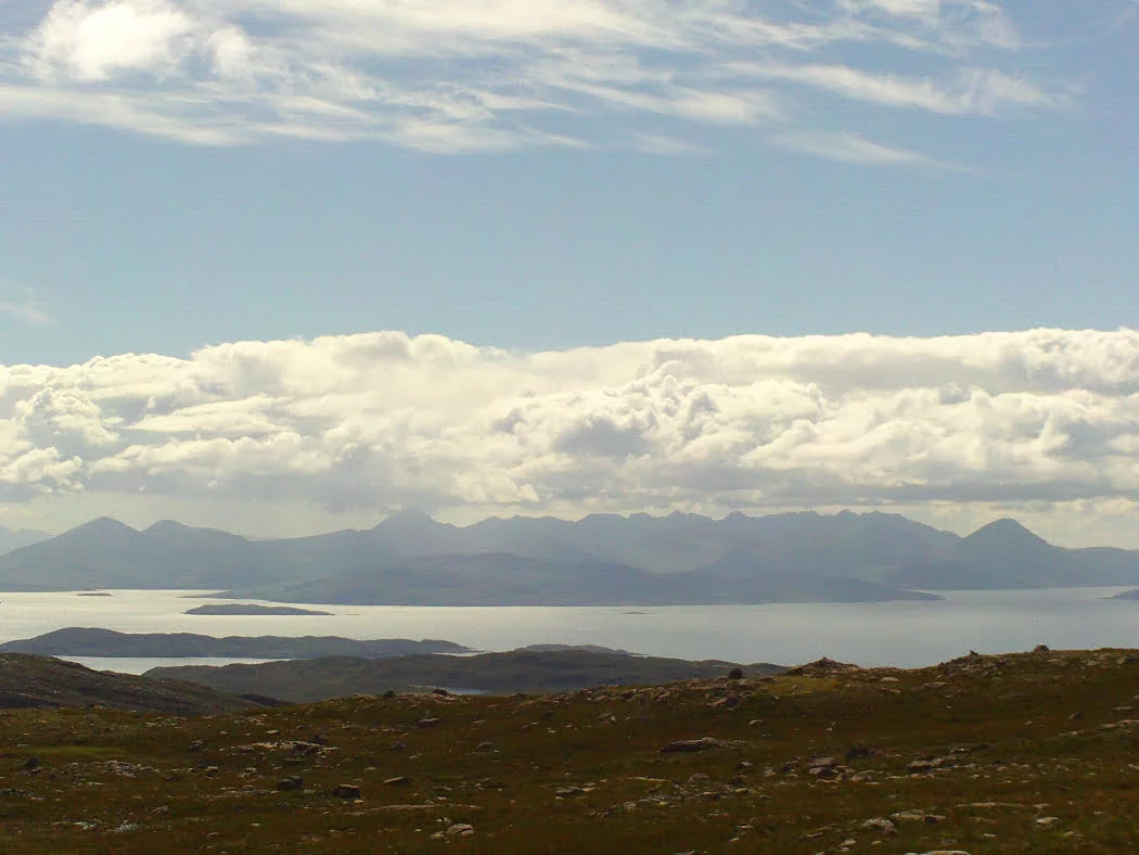 This is the view from the top of the NC500 Applecross road, which we visit along with Eilean Donan Castle. From the top of the pass rough boulder-littered ground slopes down towards the sea, two thousand feet below. The sea is a silver blue channel dotted with the silhouettes of small islands, and in the background the many blue peaks of Skye rear towards a great bank of fluffy white clouds above which all is blue. ,