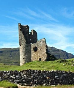 The foreground is a dry-stone wall. On a grassy knoll behind the wall stand the ruins of Ardvreck Castle. Behind that is a distant, rugged brown mountain. Above is a blue sky.