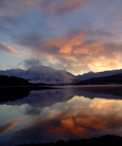A snow covered Ben Nevis is in the background while we face east , on the Fort William and the Scenic Glens Tour. Still waters of a sea inlet fill the foreground and the mountain is reflected there. There are dark coniferous forests along the shores to left and right, and the sky above is a dark blue, but lightening by sunrise near the mountain. The clouds are being lit in orange colours.