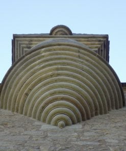 We are standing beneath a tiny turret, made from carved yellow stone. The underside, which is above our heads, is decoratively worked with delicate semi circles increasing in size as it goes upwards. That is where it meets a square stone, also worked with increasing decorative square trim. The sky is cloudless and the palest of blues.