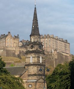 Edinburgh Castle on the Outlander Film Locations Tour is high in the background under a blue-grey sky. In the foreground is a church steeple from bottom to top of the image. to left and right of the steeple are large deciduous trees.