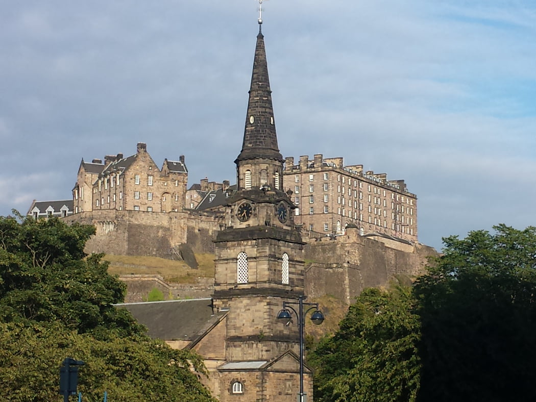 Edinburgh Castle on the Outlander Film Locations Tour is high in the background under a blue-grey sky. In the foreground is a church steeple from bottom to top of the image. to left and right of the steeple are large deciduous trees.