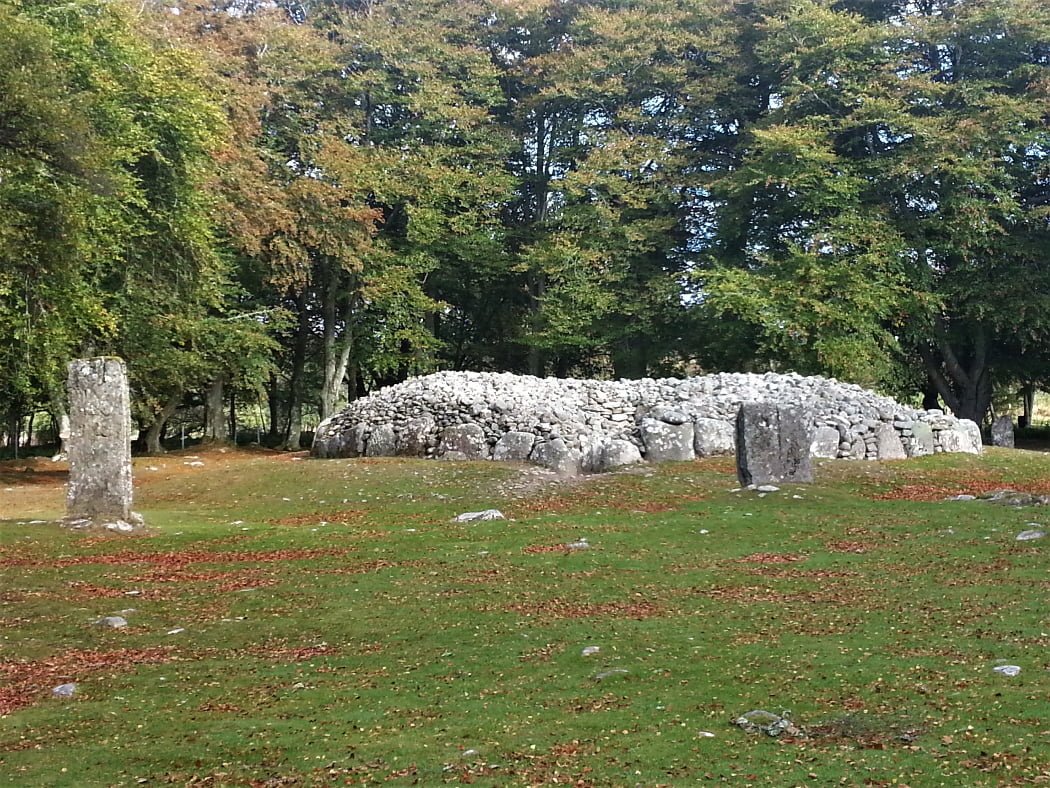 In front of us is a Clava Cairns bronze age passage grave, on the Loch Ness with Culloden and Urquhart Castle Tour. There is grass in the foreground, and two prominent standing stones, one to the extreme left, the other mid-right. Between and slightly behind them is the roofless cairn, with the mouth of the entry passage towards us. In the background are large, leafy, deciduous trees.