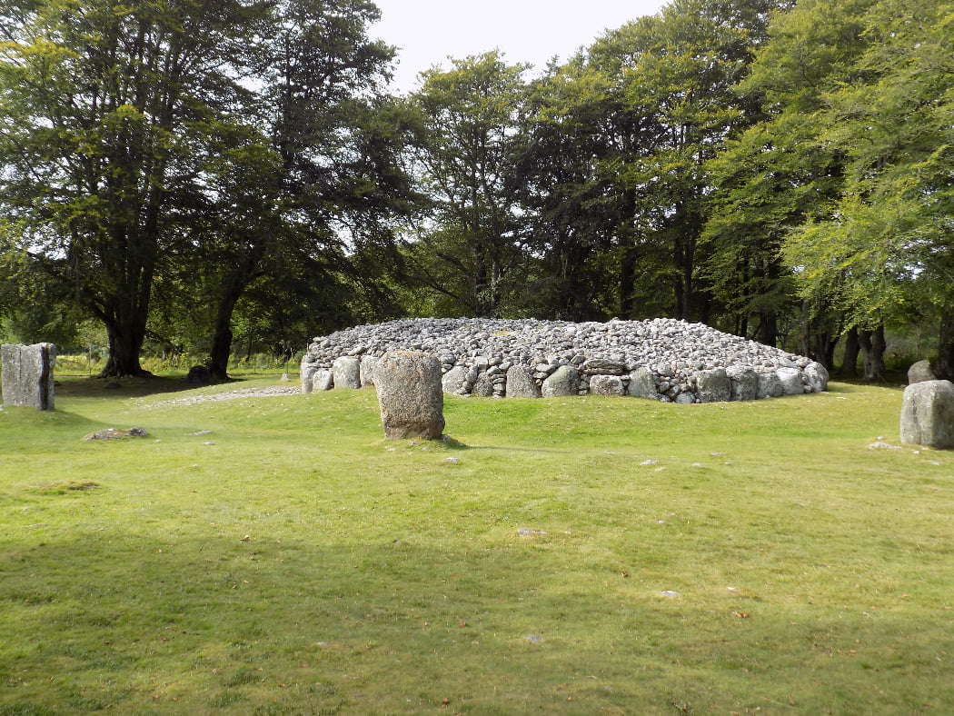 There is grass in the foreground and mid-image is the Cairn and surrounding ring of Stones on our Outlander Tour from Invergordon. Mature trees stand behind and the sky is white.