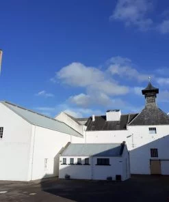 A white painted Scottish whisky distillery, Dallas Dhu, on the Ballindalloch Castle and Speyside Tour. There is a large square brick built chimney to the left, and a building with distillery-style pagoda roof to the right. The sky above is mostly blue, and with some fluffy white clouds.