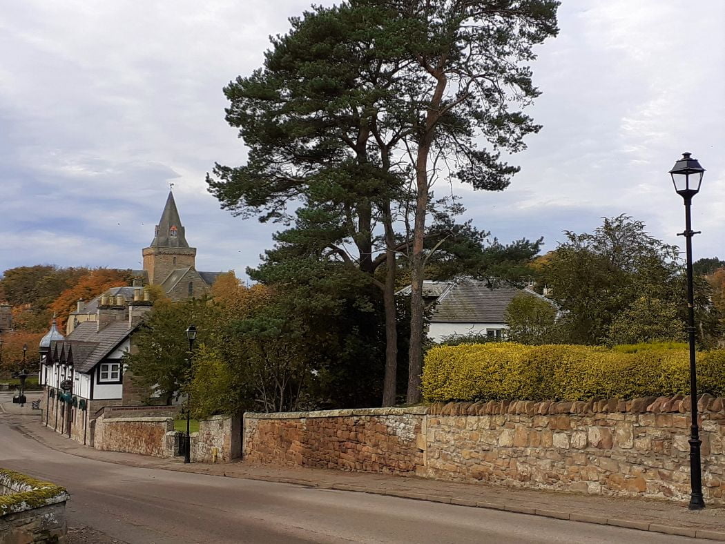 A street falls away before us, on the Rosamunde Pilcher Winter Solstice Tour. A half-timbered old building stands bottom right, and beyond it the squat square tower of Dornoch Cathedral. to the right is an old black lamp-post. The Skye above is blue and cloud.