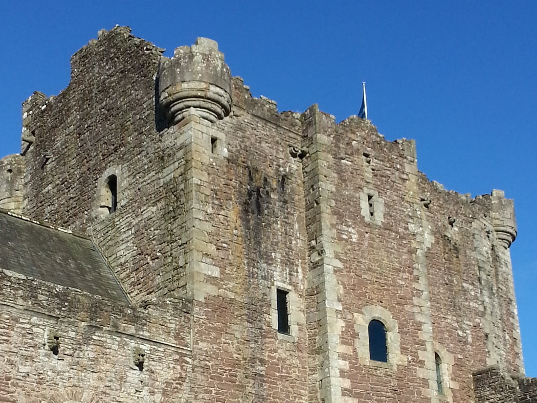 The grey keep of Doune Castle rears into a cloudless blue sky, on the Highland and Lowland Distillery Tour. There is a flagpole centrally sited on the battlements of the keep, flying the Scottish flag.