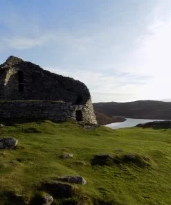 A ruined iron age tower called Dun Carloway, on a rocky green hill under a sunny blue sky, with a sea inlet behind. This is visited during both our Hebrides Tours