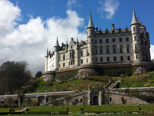 A huge chateau-style castle dominates this image, standing above us on a rise. Many-turreted and white, it stands under a blue, but cloudy, sky and gardens are at the castle's feet.