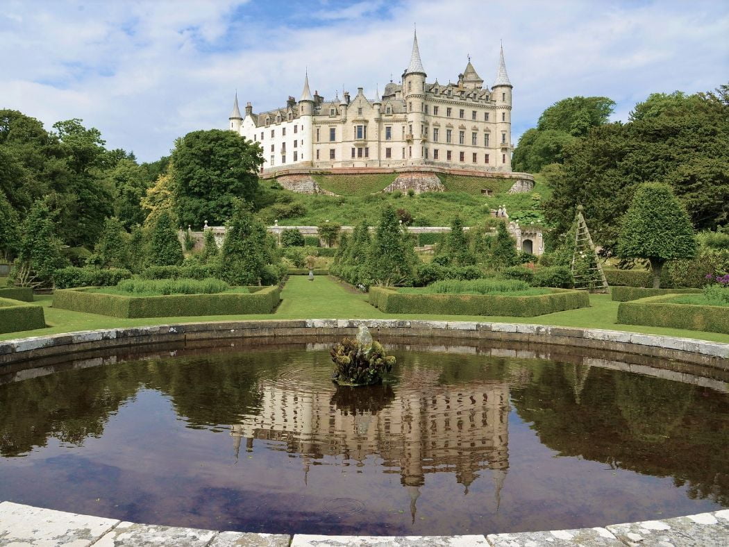 Ornate gardens and oval pond in the foreground, with retaining wall in the middle distance. Above this the gardens rise steeply, terminating in the major stone built foundations of a vast white Jacobean Chateau rising four storeys into a blue sky and multiple fairytale conical towers.