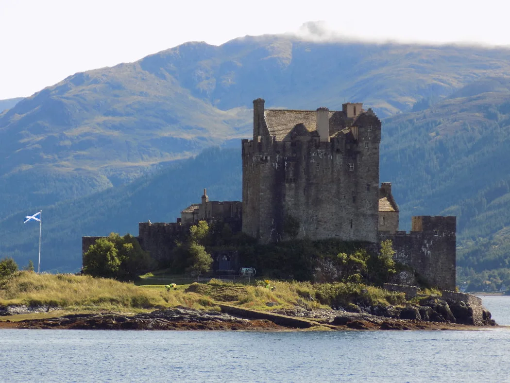 Eilean Donan Castle on the NC500 Applecross tour is across a very narrow arm of the sea. We are nearly at sea level, and a couple of hundred yards away it is built of grey stone rising from a grassy, rocky outcrop. A Scottish Flag flies on a ground mounted flagpole to the left of the castle. A large mountain clothed in coniferous forest rises behind the castle.
