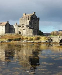 The main area of this picture is the grey Keep of Eilean Donan Castle on an islet, and the arched stone bridge connected it to the mainland exits the frame on the right. The foreground is the slightly rippled reflection of the castle in the sea. The sky above is dark and cloudy.