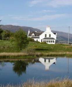 A picturesque traditional white painted building with the word hotel painted on its gable. It stands on grass, and has trees to the left, behind it. The sky is blue and the building is perfectly reflected in a blue pond in the foreground.