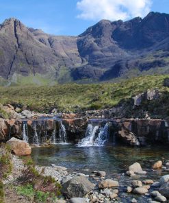 The Fairy Pools, which can be visited on our Isle of Skye Three Day Tour, are shown in this picture. A small river makes a low white-water cascade into a clear pool. Behind are the rocky peaks of the Black Cuillin Mountains. The sky is blue with puffy white clouds.