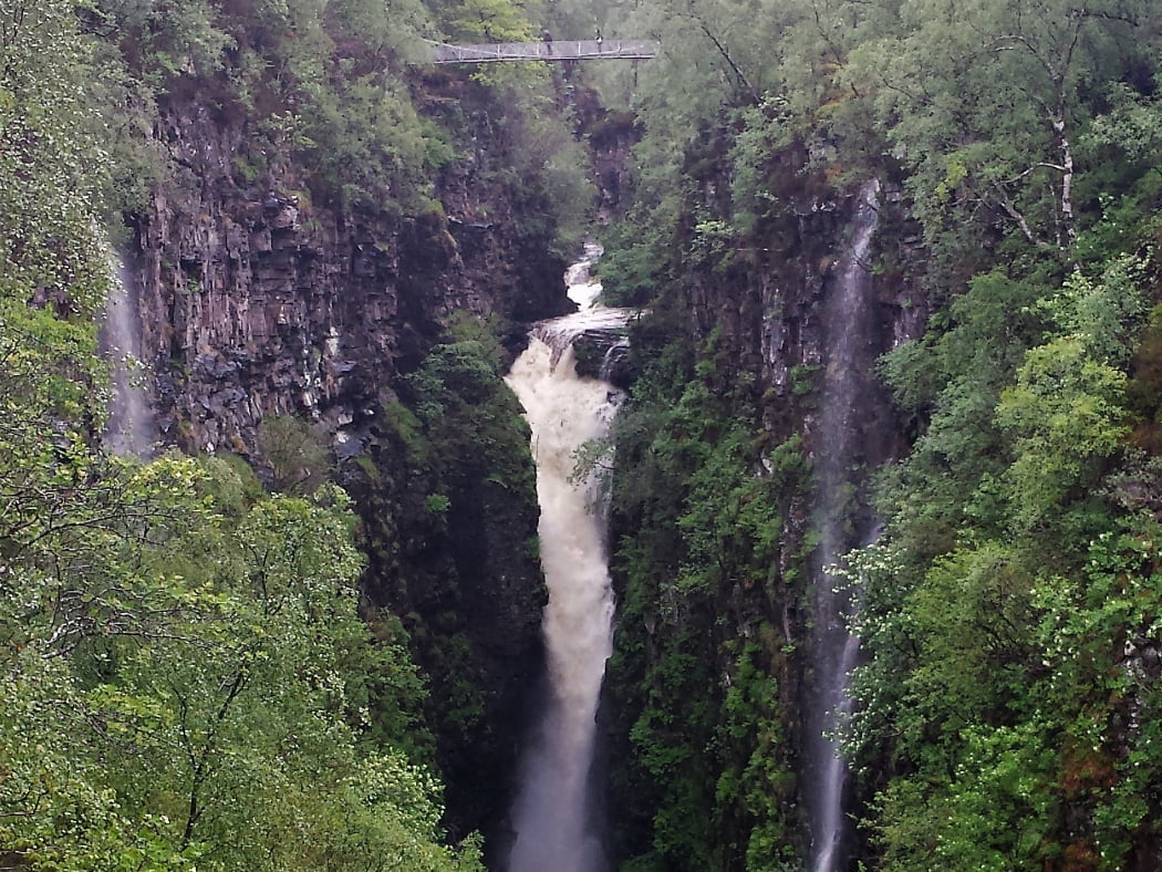 Huge waterfall falling into a gorge on our Inverewe Gardens and Waterfalls Tour. The canyon sides are tree clad and moss covered. Two smaller falls can be seen falling into the gorge (one on each side). A footbridge spans the main falls and no sky is visible.