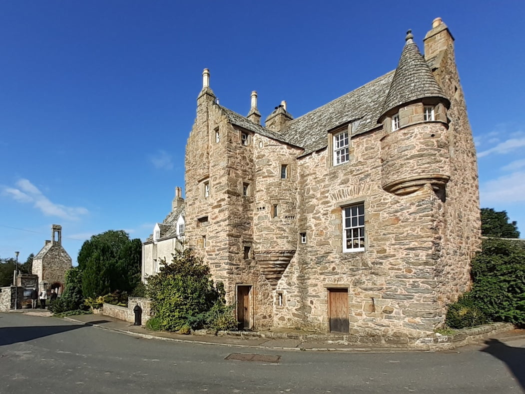 Fordyce Castle, a rustic stone-built Scottish towerhouse fills most of the picture, standing on the village street. To the left is the ancient churchyard beneath a deep blue sky.