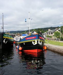 Two brightly painted long canal barges in the waters of a lock gate at Fort Augustus on the Loch Ness and Urquhart Castle Tour. Old village houses can be seen on both banks of the canal, and there are three flags flying outside a house on the right. A man in lifejacket is coiling the rope from the right hand barge around a hook on the ground.