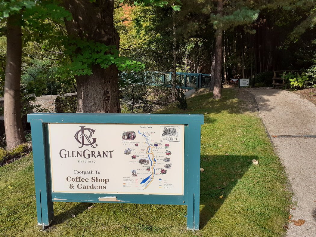 The main item in this image taken on our Speyside Distillery Tour from Inverness is a timber sign that simply says Glen Grant, footpath to Coffee Shop & Gardens Distillery and has a small map. A gravel path runs up the left side of the image, and there is a bit of grass behind the sign and lots of trees.