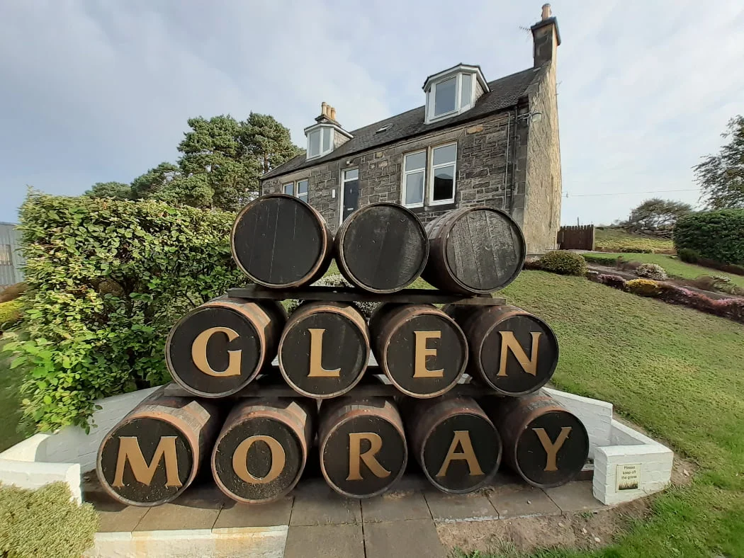 A barrel stack is in front of us. The barrels are on their sides. The top row has three barrels, plain ends. The middle row has four barrels each with a letter on it. The bottom row has five barrels each with a letter on it. The letters spell the words Glen Moray. Behind the barrel stack is a stone built house of two storeys. A door is centrally placed on the ground floor, with a window either side, and two dormer windows above.