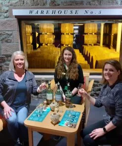 Three women sit at a table, with glasses and three bottles of whisky. Two are in the act of toasting, and the one at the back of the table (in the middle) has no glass and appears to be a badged guide. Behind them is a stone wall with a window, giving a view into a lit whisky-barrel warehouse.