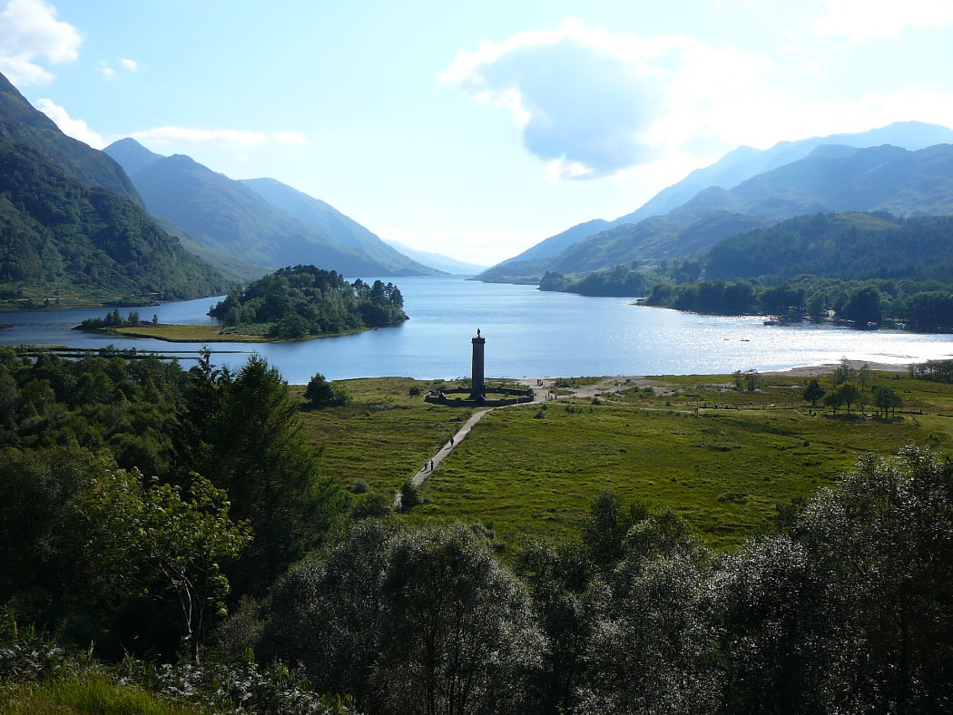 Fort William and the Scenic Glens Tour visits Glenfinnan, the slender stone tower with statue of kilted Highlander on top. Behind the tower a narrow lake disappears into the distance between tall mountains clothed in some native woodland.