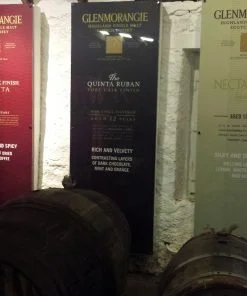 There are three 6 foot tall advertising boards which each describe a different whisky from Glenmorangie Distillery on our Highland Distillery Tour from Inverness. We are inside a warehouse, and in the foreground, on their sides are three barrels, one at the foot of each advertising banner.