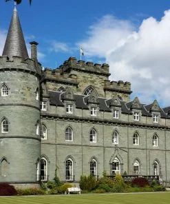 Inveraray Castle on our Loch Lomond and Inveraray Castle Tour dominates and fills this shot. A gothic revival castle built in the 1700's, it is grey, square and with four round corner towers with conical roofs. Green lawns are at the foot of the picture and a cloudy blue sky above.