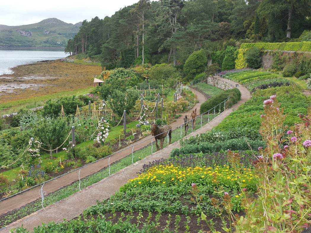 A colourful walled garden on the shore, with a pine clad headland behind and the sea to the left. A wicker sculpture of a horse and two humans makes an interesting garden centrepiece.Inverewe Gardens - Inverewe Gardens and Waterfalls Tour