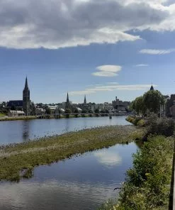 The River Ness is on the left, flowing towards us. On the opposite bank is a street of old houses, and 4 different steepled buildings, interspersed at distances upstream. The most distant building on the far bank is Inverness Castle, against the sky. The day is sunny and the sky is mainly blue. On our bank of the river are a line of mature deciduous trees, in leaf, and another street of old houses. There is a shingle bar to our immediate left, in the river.