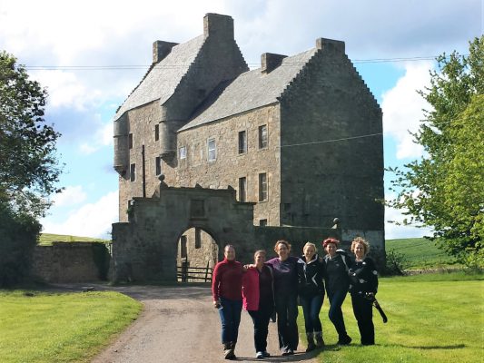 A gravel road with grass each side leads to the 5-storey grey stone towerhouse in the centre of the picture. Six ladies stand in the sun,in a line on road and grass in front of the towerhouse. There are trees to either side. The sky is cloudy and light blue.