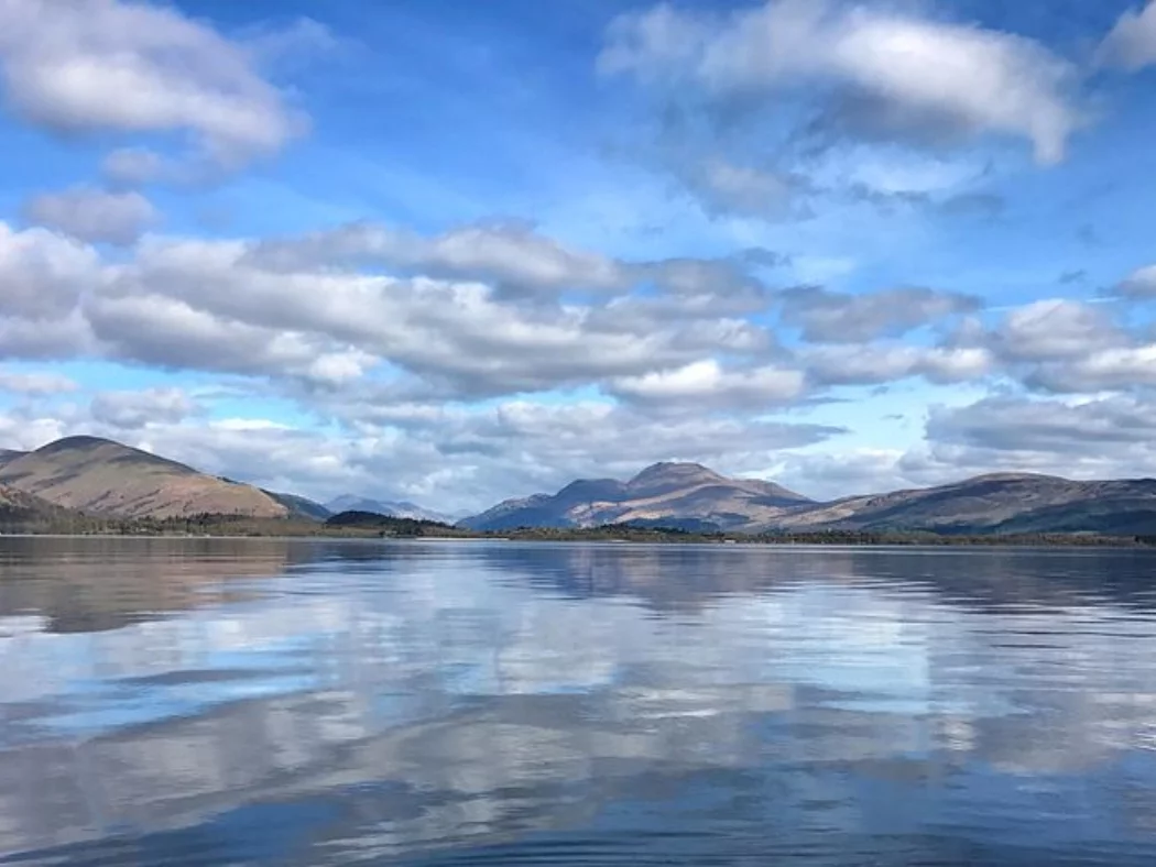 The lower half of this image is Loch Lomond on our Stirling Castle and Highland Cattle Tour. The far shore of the loch comprises the many mountains of the Highlands, blue and blue-green in colour. The sky is filled with clouds and is a mix of blue and white.