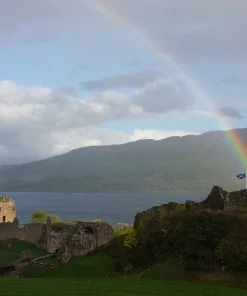 A ruined castle castle on a rock in a grey-blue lake. The tower at the left end is sunlit, but the rest of the ruins not. Except there is a vivid rainbow arcing straight down upon the Scottish flag flying at the castle highpoint.