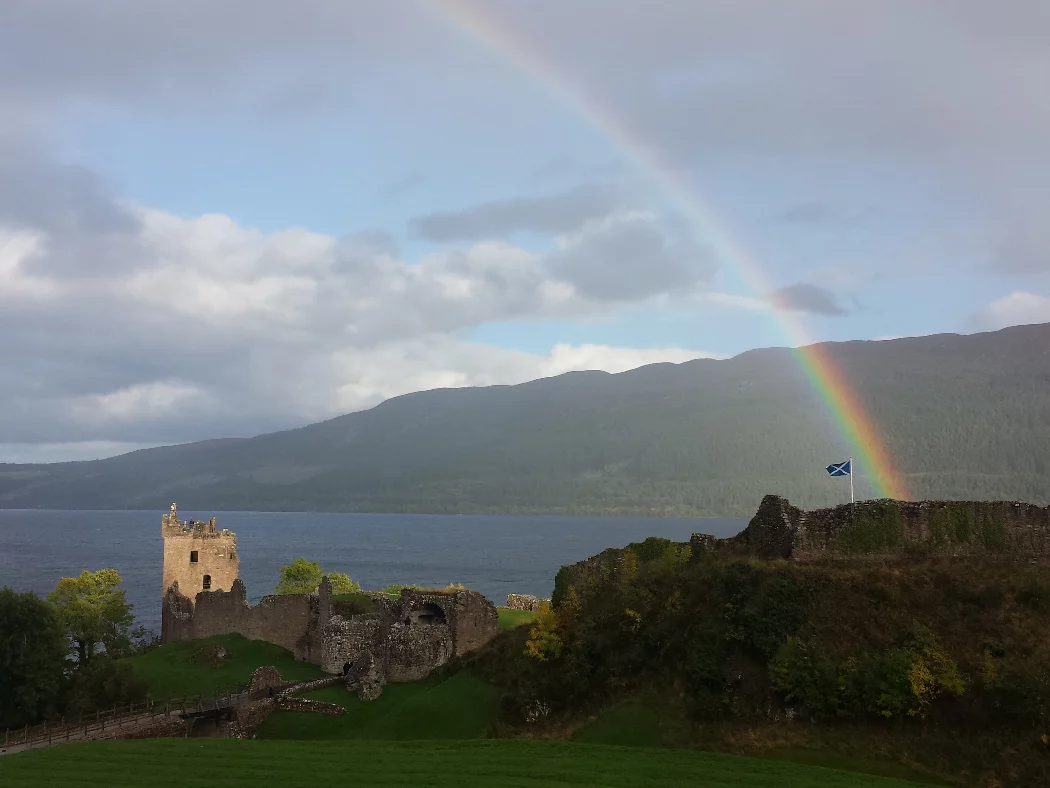 A ruined castle castle on a rock in a grey-blue lake. The tower at the left end is sunlit, but the rest of the ruins not. Except there is a vivid rainbow arcing straight down upon the Scottish flag flying at the castle highpoint.