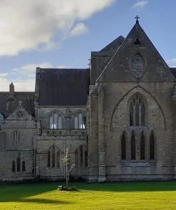The foreground is a beautifully mown lawn, and most of the rest of the frame is filled with the main buildings of a restored 1200's abbey, in grey stone with slate roofs. The sky is cloudy blue.
