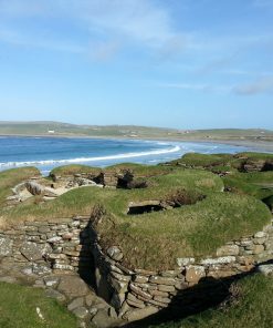 In the foreground are the low ruins of Skara Brae, Stone Age village, visited on our Orkney Tours. Behind this is the broad crescent of the Bay Of Skaill, with a lazy Atlantic swell coming in over the sand, under a blue sky.