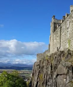 A close-up view of Stirling Castle, on our Wallace Monument Tour fills the right side of the image. The castle is above the line of sight, and the cliff falls from our location. The day is fine and across the flat green fields below us, we can see the blue mountains of the Highlands on the far horizon.