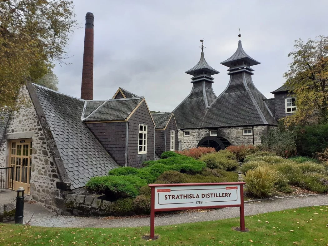 1700's Strathisla Distillery on our Moray Coast and Speyside Tour. In the foreground the wide distillery sign stands on grass, and behind is a tall chimney, two pagoda roofed drying rooms and a water wheel. There are trees in autumn colours to left and right and the sky is grey.