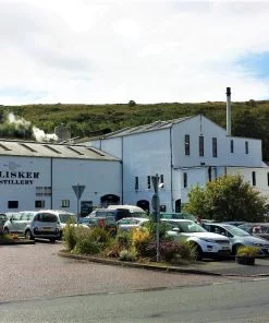 In the foreground is a full car park. The large white painted building occupying the whole of the middle-distance says Talisker Distillery on it. In the background a green hill rises towards a mixed blue and cloudy sky.