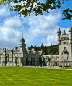 Balmoral Castle fills this picture. There are green lawns in front of the castle and the Royal Standard flies from the highest tower. There are forested hills behind, and a cloudy and blue sky above.
