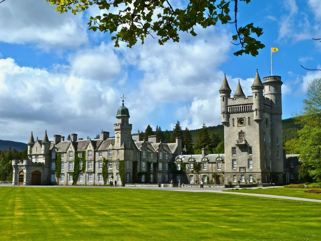 Balmoral Castle fills this picture. There are green lawns in front of the castle and the Royal Standard flies from the highest tower. There are forested hills behind, and a cloudy and blue sky above.
