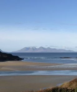 Sandy beach with wooded headlands to right and left. The blue sea is in the background and blue peaked islands lie out on the horizon under a blue sky.