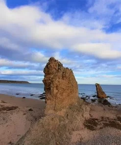 Two red coloured rock pinnacles rising from a sandy beach. The ocean is behind them and the sky is a mix of cloud and blue.