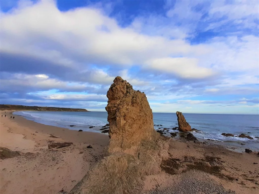 Two red coloured rock pinnacles rising from a sandy beach. The ocean is behind them and the sky is a mix of cloud and blue.