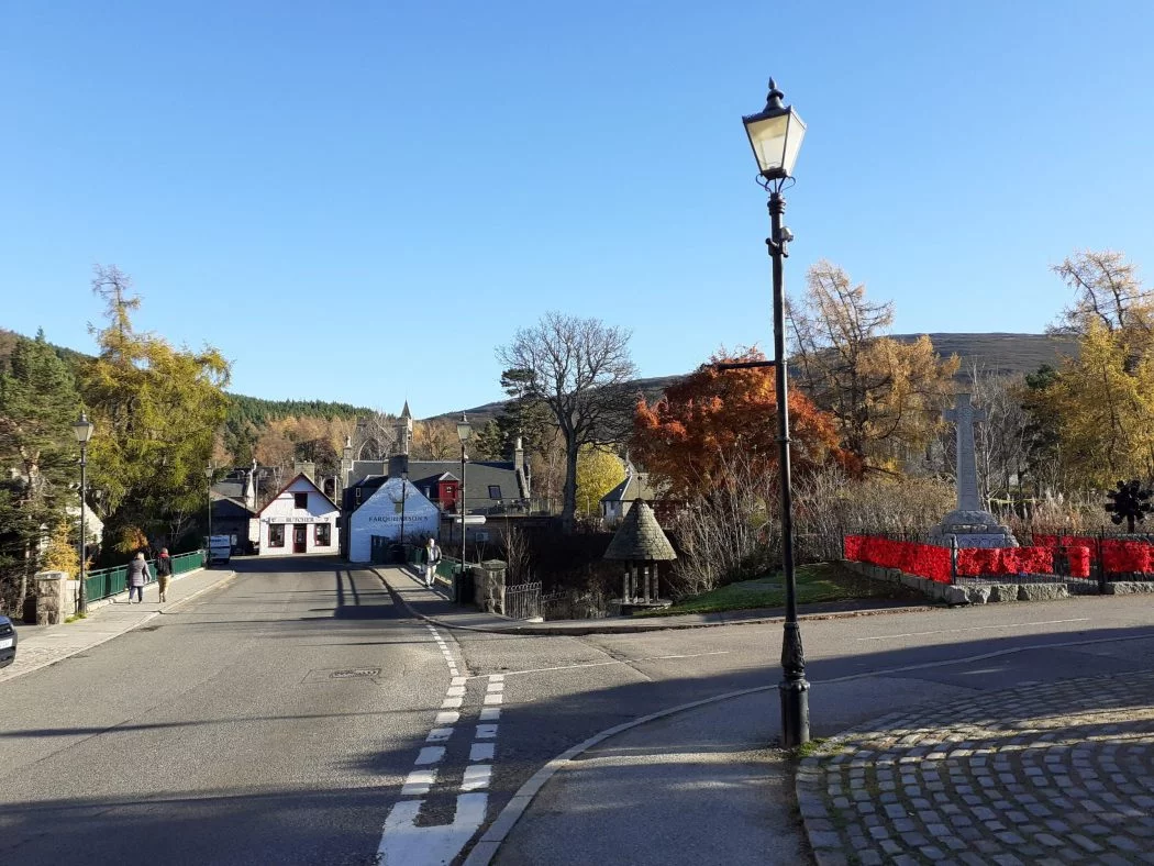 The town of Braemar on the Balmoral Castle and Royal Deeside Tour. The sky is cloudless blue, and we stand on the roadside by a historic black-painted streetlamp. There is a bridge in the foreground and at the other end of it are quaint whitewashed old buildings containing a shop and a pub.