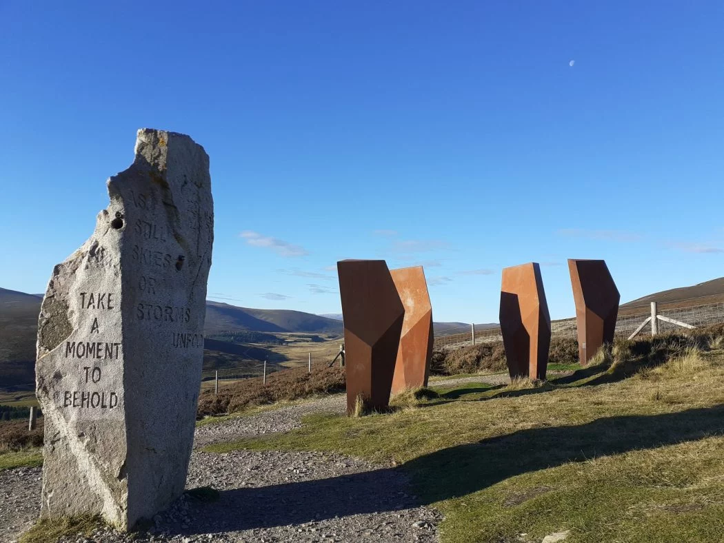 Five modern Standing Stones, four of which are geometric. They stand in grass and gravel on a hillside above a valley. The sky is blue, and a winter moon hangs above this art installation.