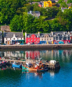 Arial view of Tobermory on our Isle of Skye and Mull Tour. Colourful houses line the harbourfront and a stone built pier projects into a calm blue sea. Six colourful fishing boats are tied to the pier.