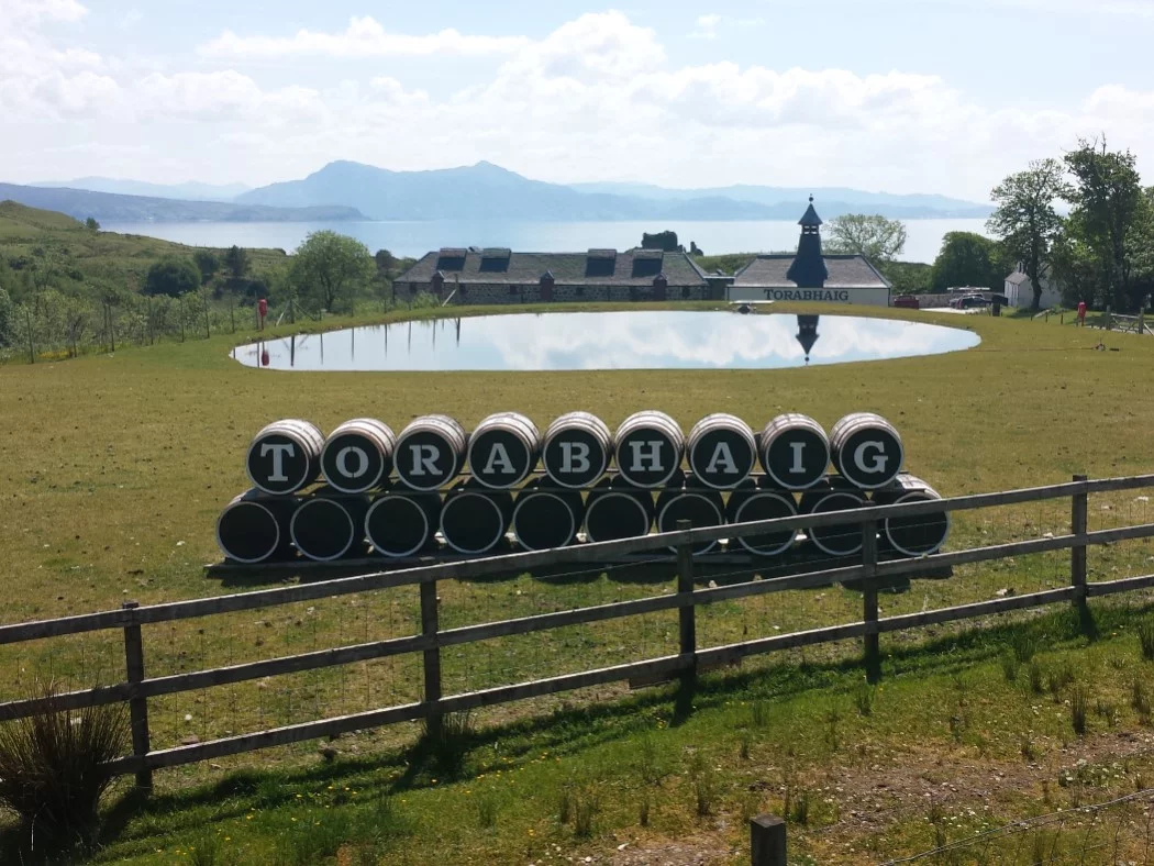 In the background are the faded blue mountains of the mainland and in the foreground is the new distillery, Torobhaig. We sometimes visit it on the Isle of Skye and Hogwarts Express Tour. It is a long low building above the sea, and it is reflected in a pond on the grass in front of us. Closest to us is a line of barrels. Each has a letter painted on it, spelling out the name Torabhaig.