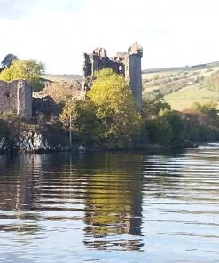 A ruined castle rises on a wooded outcrop from the waters of a lake. Our viewpoint is from a boat offshore. In the background, behind the castle rises a hill with farms and farmland upon it. The sky is white.